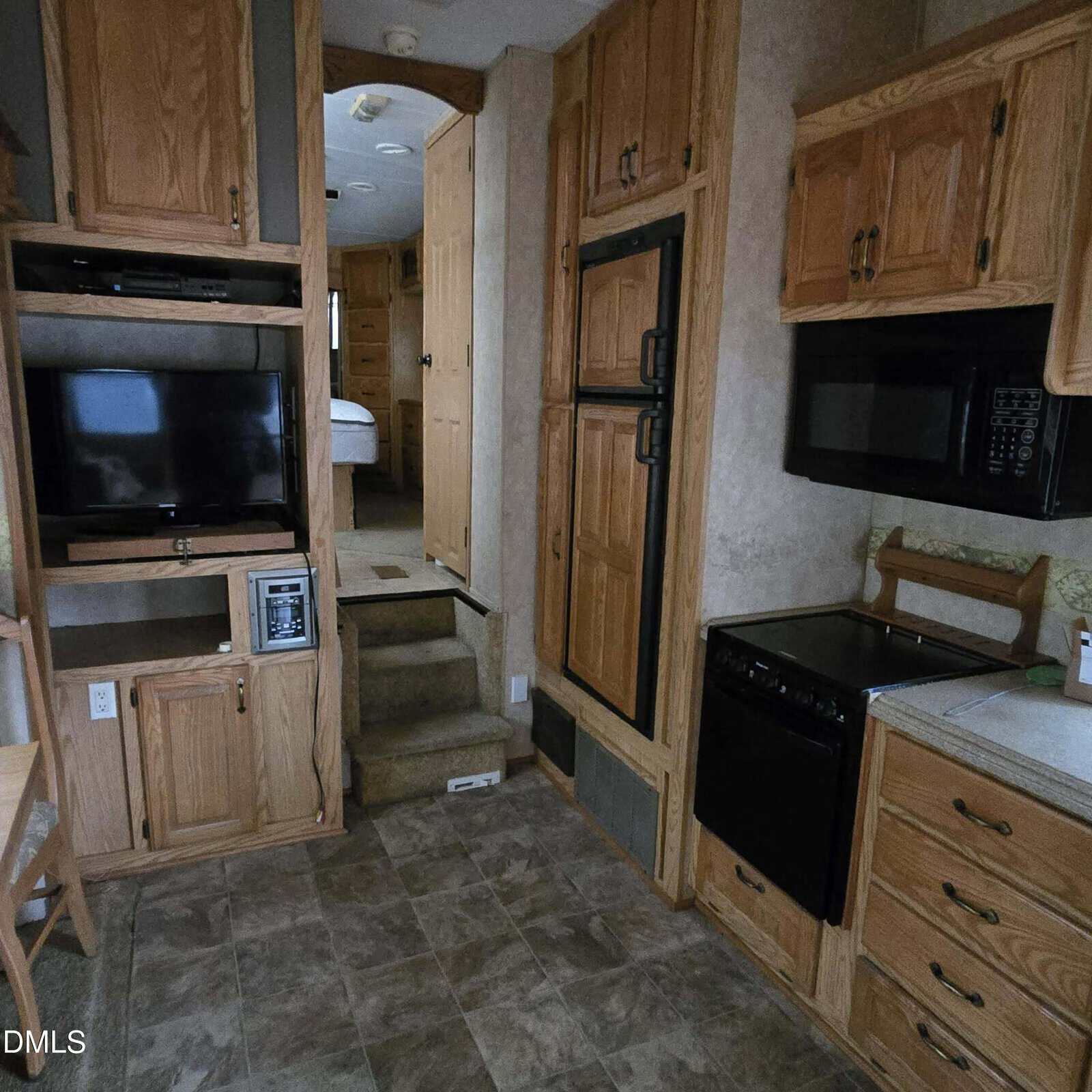 Interior of an RV kitchen with wooden cabinets, TV, microwave, stove, refrigerator, and small stairs leading to a raised bedroom area with bed visible in the background.