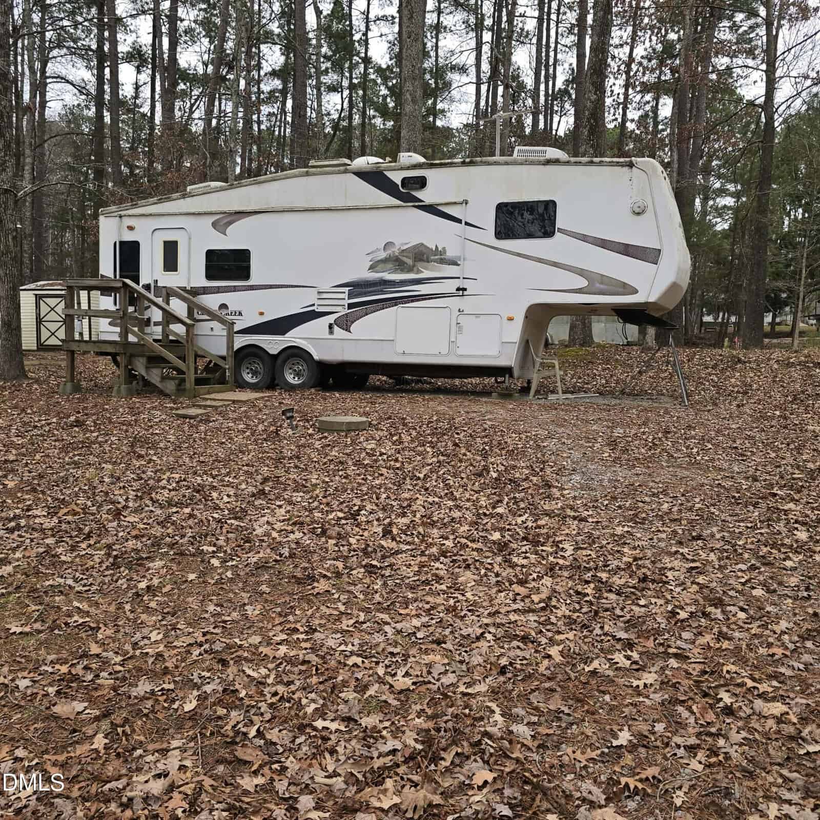 A white fifth-wheel RV with a mountain-themed decal is parked among tall trees on a lot covered with dry brown leaves. A small wooden staircase leads to the RV entrance.