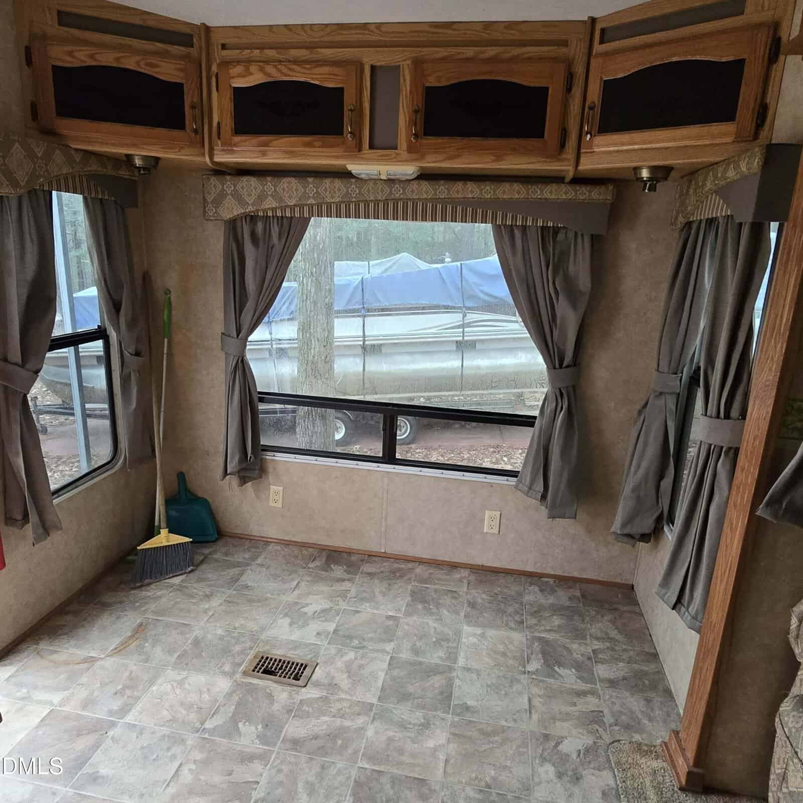 Empty RV interior with tile-patterned floor, two gray curtained windows, wooden overhead cabinets, and a broom and dustpan in the corner. A boat is visible outside through the window.