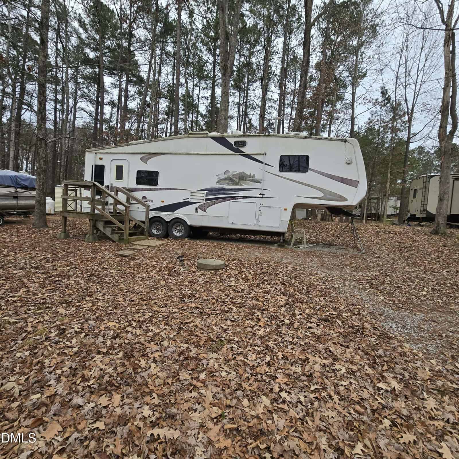 A white fifth-wheel camper with mountain graphics is parked among tall trees at 113 Red Wing Drive, Louisburg, surrounded by fallen leaves. Wooden steps lead to the camper door, and other campers are visible nearby.