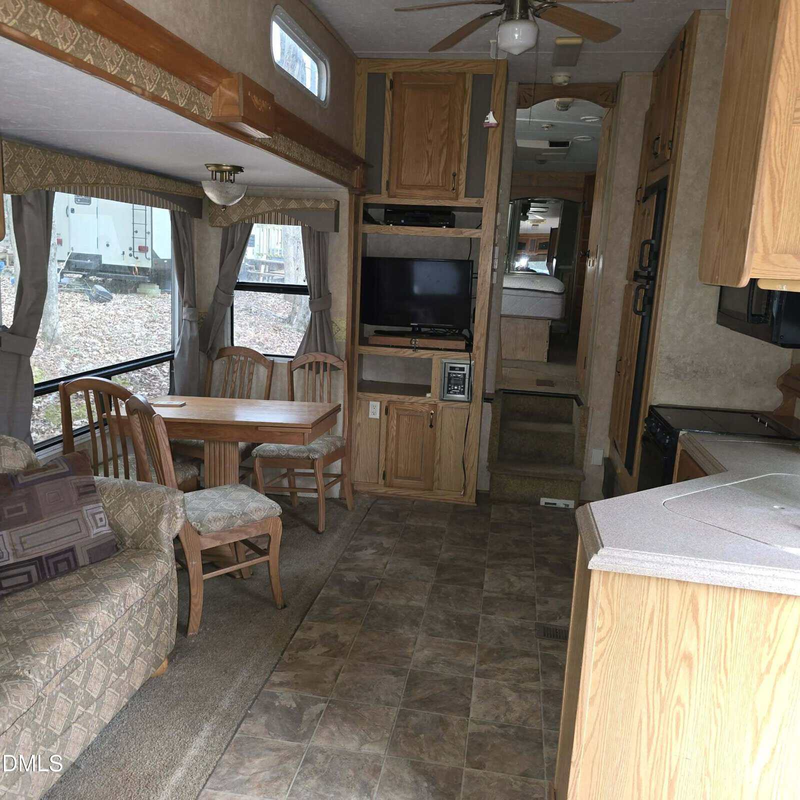 Interior of an RV with tiled flooring, a small dining table with four chairs, a cushioned sofa, built-in TV, wooden cabinets, kitchen counter, and stairs leading to a raised bedroom area in the back.