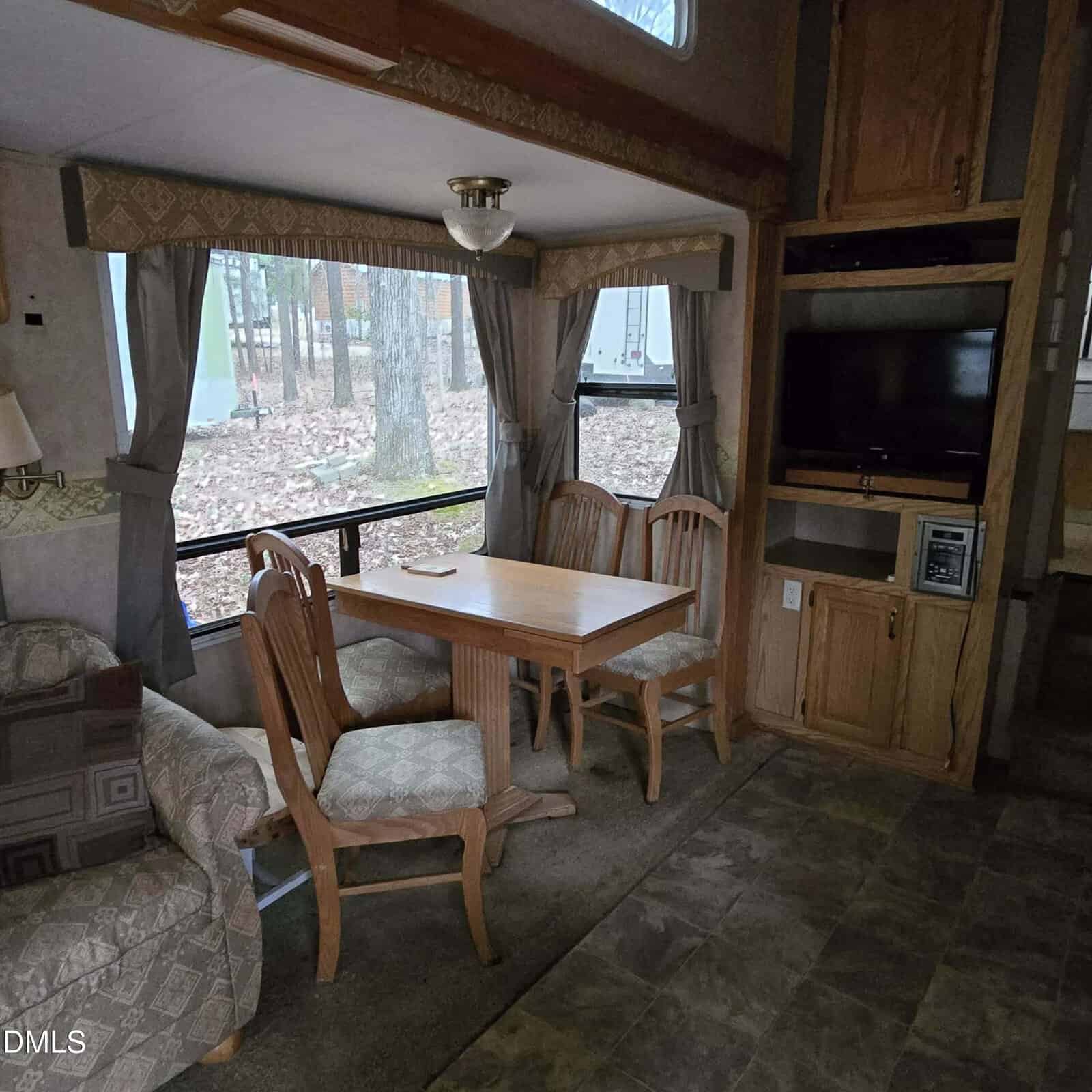 A cozy RV interior with a wooden dining table and four chairs by a large window with gray curtains, an armchair, and a built-in TV and microwave in wooden cabinetry. Trees are visible outside the window.