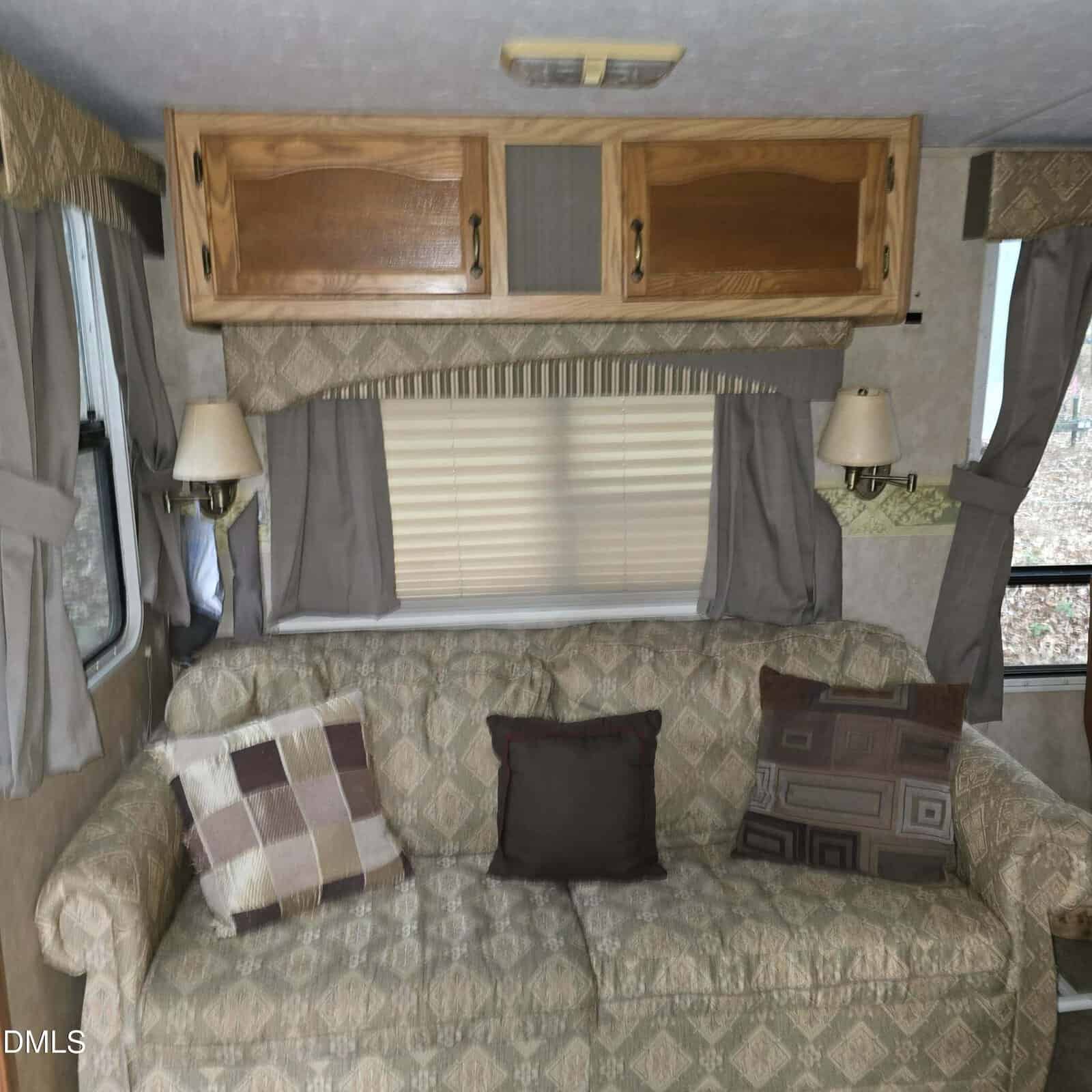 A patterned sofa with three decorative pillows sits against a wall with a window, beige blinds, gray curtains, and built-in wooden cabinets above in an RV interior. Two wall-mounted lamps are on either side of the sofa.