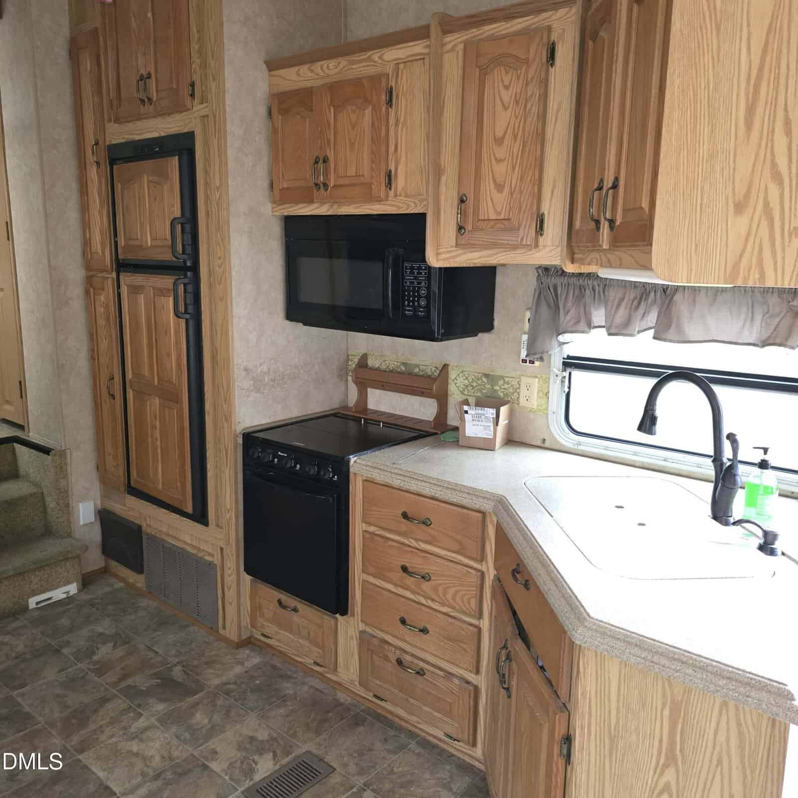 Kitchen area inside an RV featuring wood cabinets, a black stove with oven, black microwave, two-door refrigerator, L-shaped countertop with sink, faucet, cleaning supplies, and a window with a curtain above the sink.