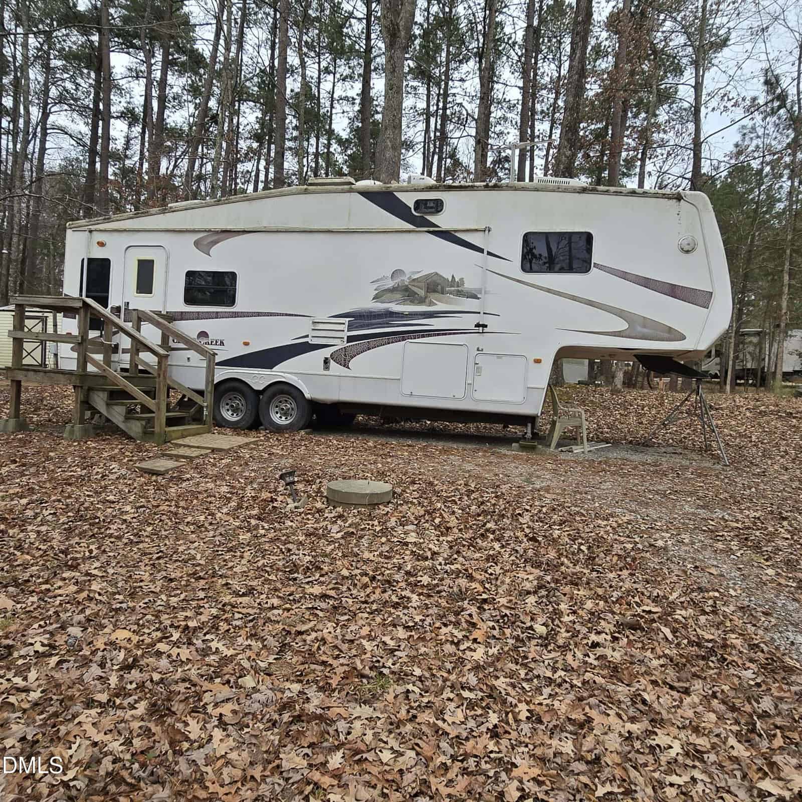 A white fifth-wheel RV trailer with mountain graphics is parked in a wooded area on fallen leaves, next to a small wooden staircase and surrounded by tall trees with brown leaves.