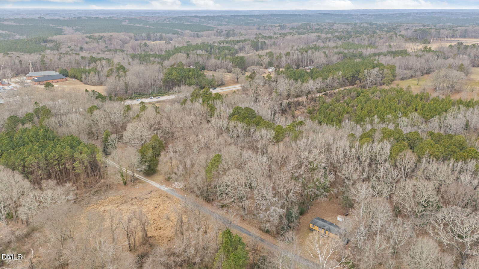 Aerial view of a rural, wooded landscape with leafless trees, scattered houses, and winding roads cutting through the forest under a cloudy sky.