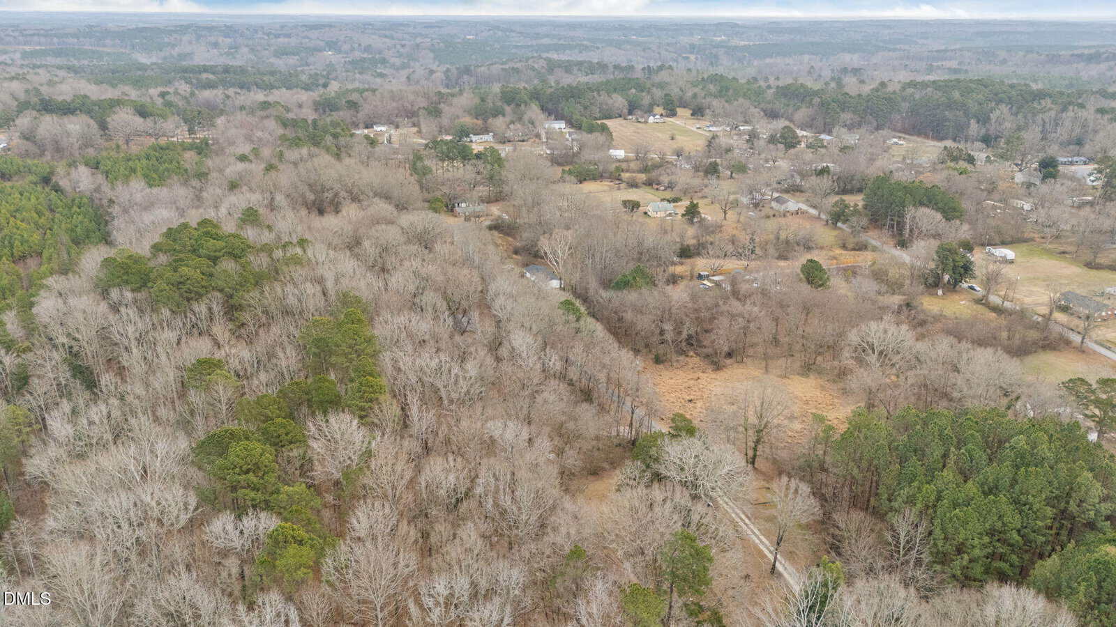 Aerial view of a rural landscape with dense woods, scattered houses, open fields, and winding roads under a cloudy sky.