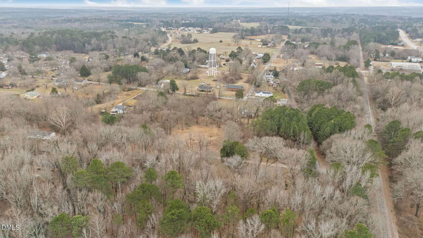 Aerial view of a small rural town with a water tower in the center, surrounded by scattered houses, trees, fields, and a winding road on a cloudy day.