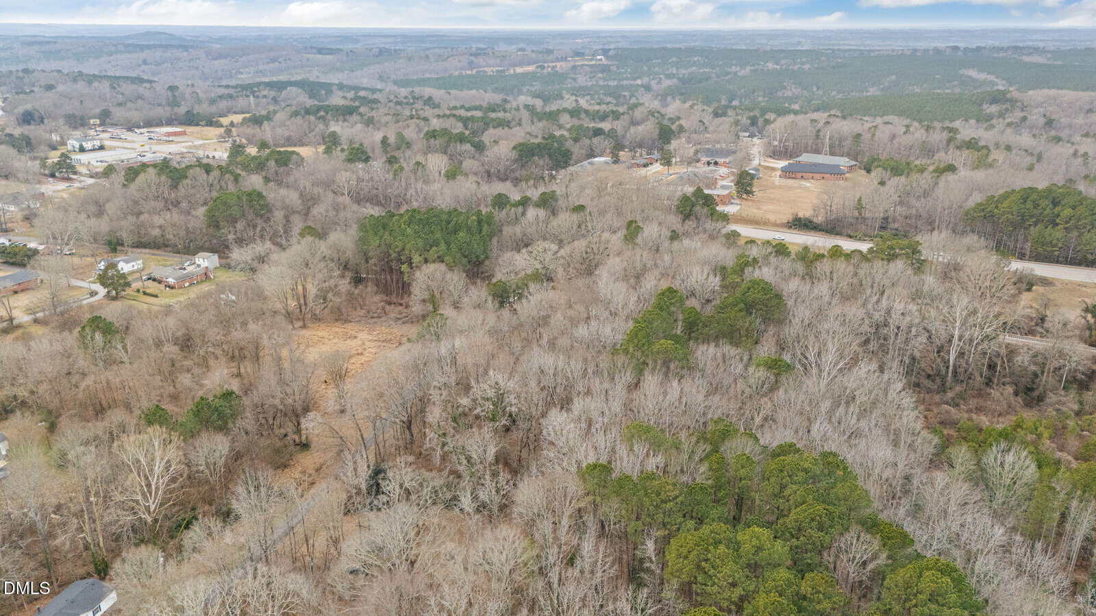 Aerial view of a rural landscape with clusters of trees, open fields, scattered houses, and a few buildings near a main road, under a partly cloudy sky.