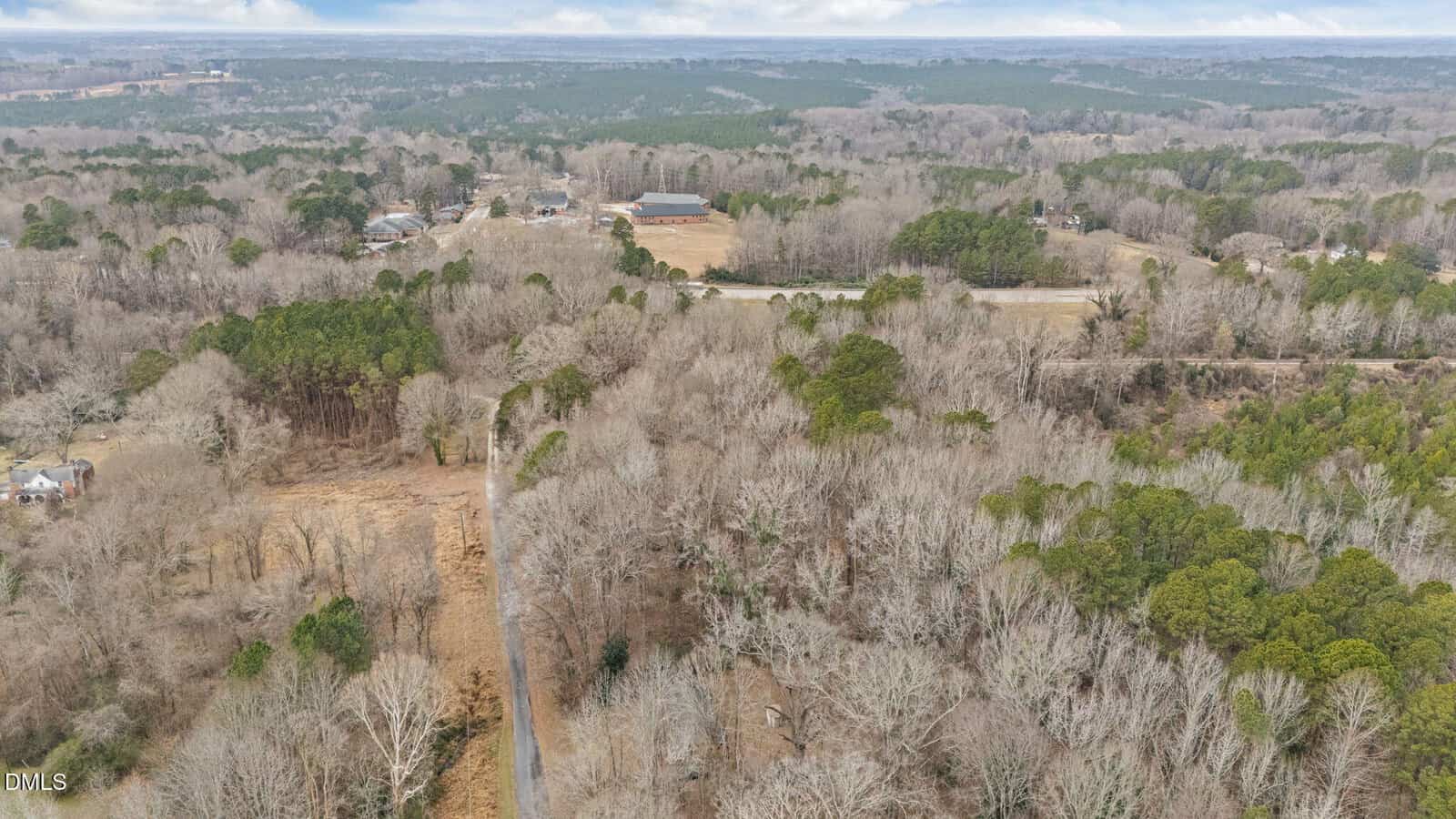 Aerial view of a rural area with a narrow road winding through leafless trees, open fields, scattered houses, and dense woods under a cloudy sky.