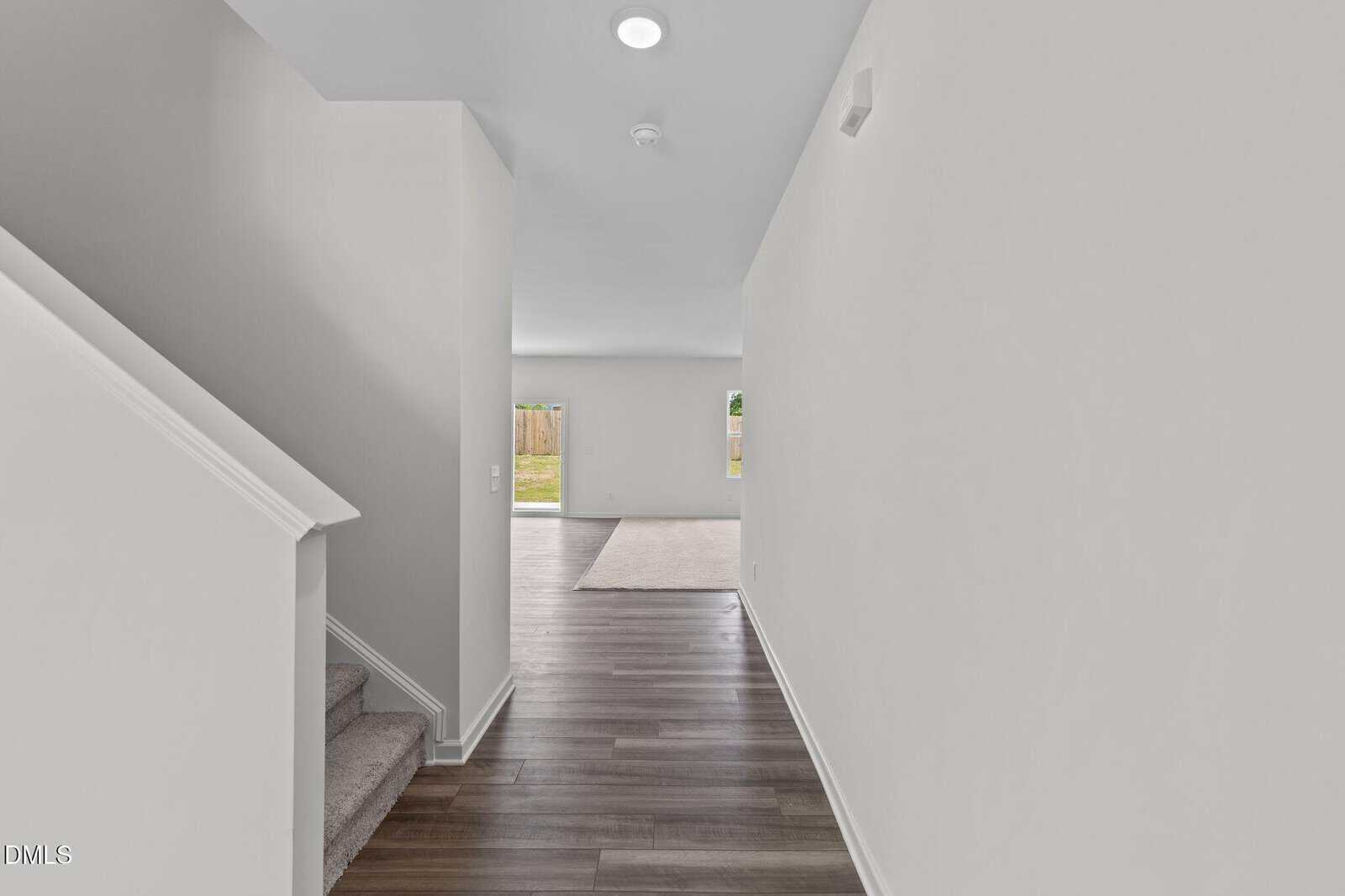 A bright hallway with wood flooring leads to a living area with carpet; there is a staircase on the left and large glass doors at the far end open to a backyard.