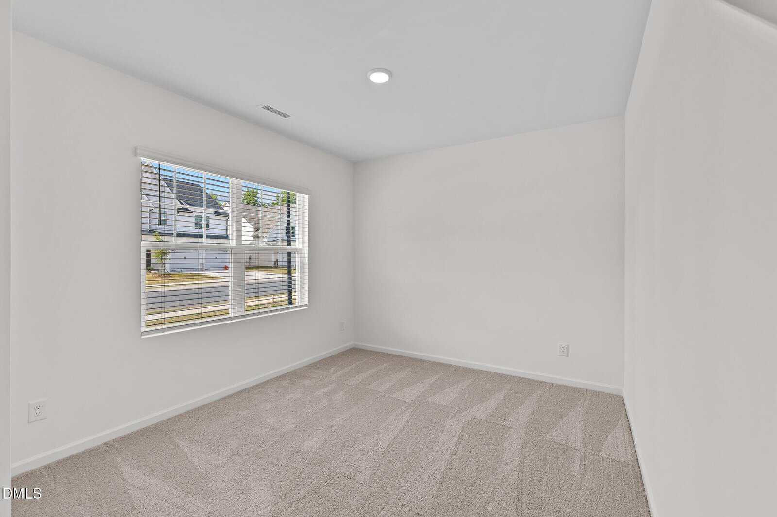A bright, empty room with white walls, beige carpet, and a large window with blinds, letting in natural light. The window shows a view of a suburban street and houses outside.
