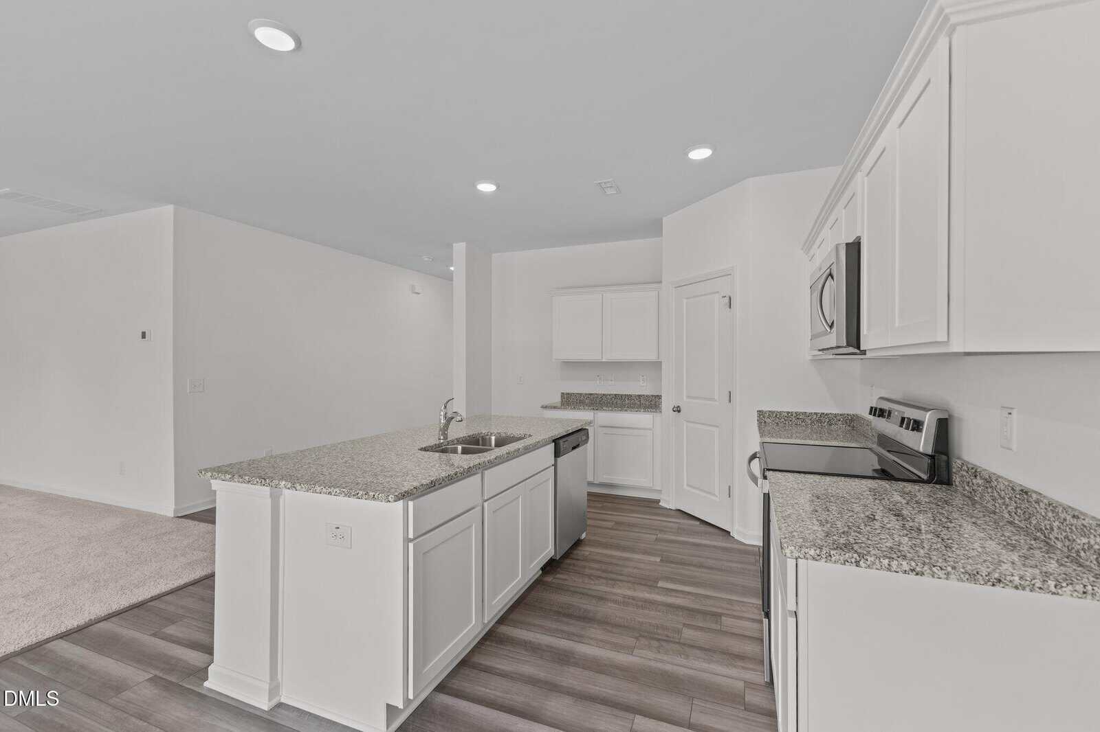 A modern kitchen with white cabinets, granite countertops, stainless steel appliances, an island with a sink, and wood-look flooring, opening into a carpeted living area. The space is bright and minimalistic.