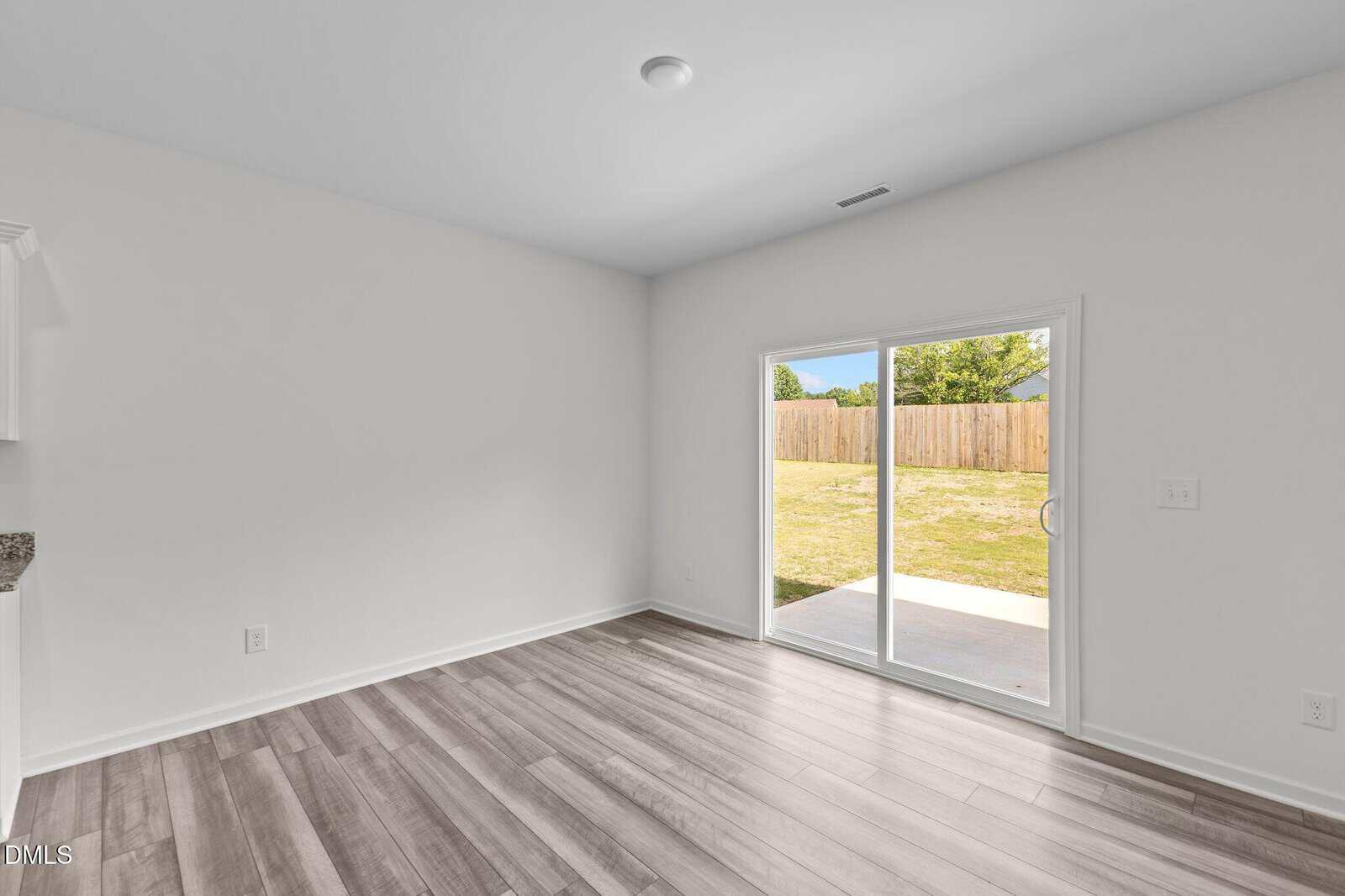 Bright, empty room with light-colored wood flooring, white walls, and a sliding glass door opening to a backyard with a wooden fence and green grass.