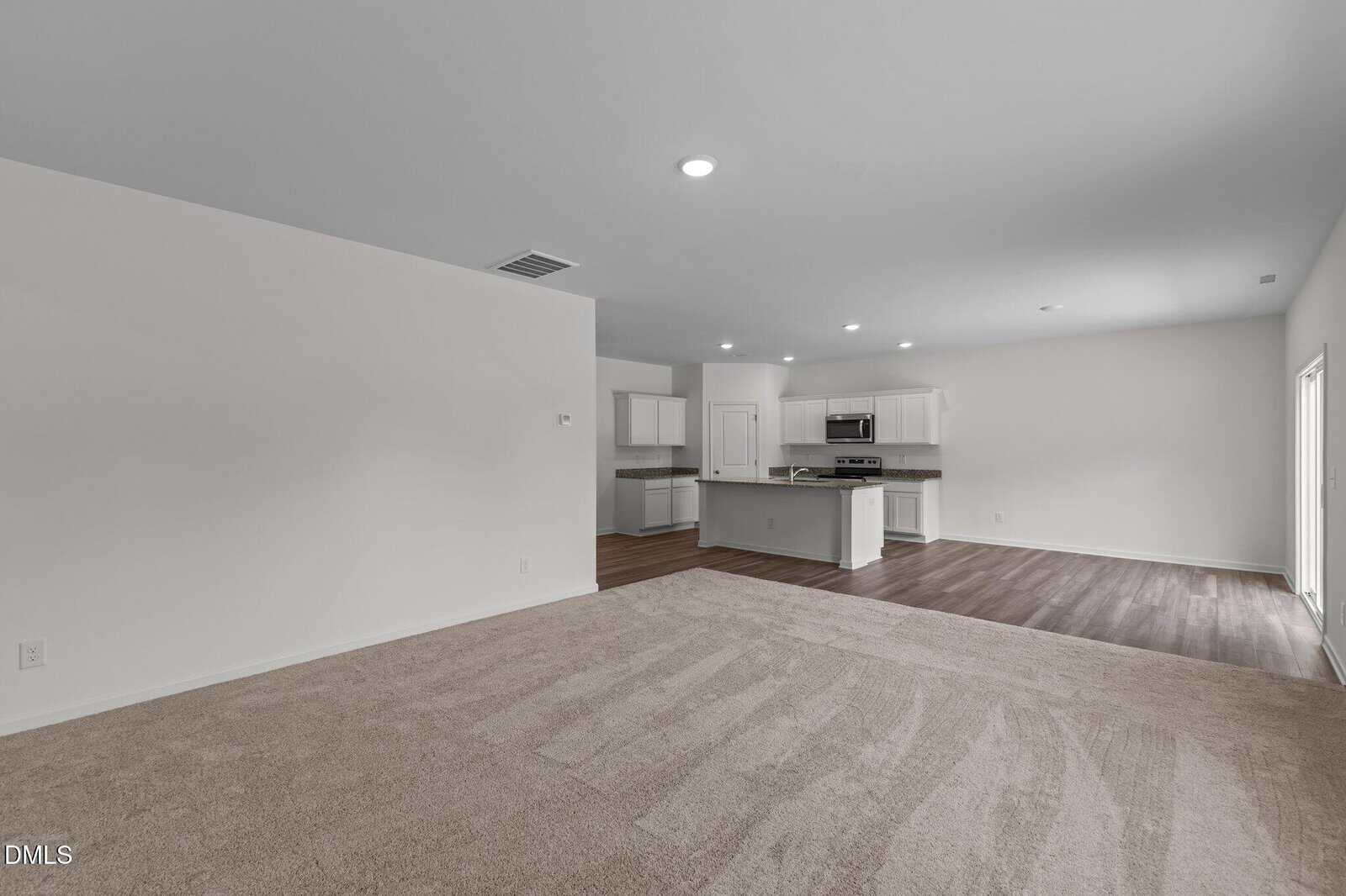 Spacious, empty open-plan room with beige carpet in the living area, wood flooring in the kitchen, white walls, recessed lighting, and modern white kitchen cabinets with stainless steel appliances.