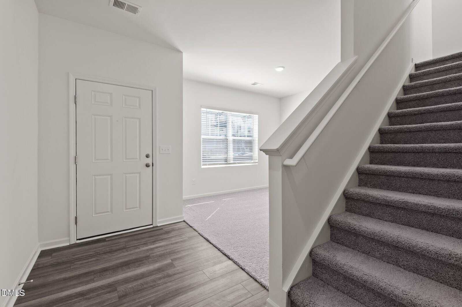 A modern home entryway with a white front door, wood-style flooring, carpeted stairs, and a bright living area with a large window letting in natural light.