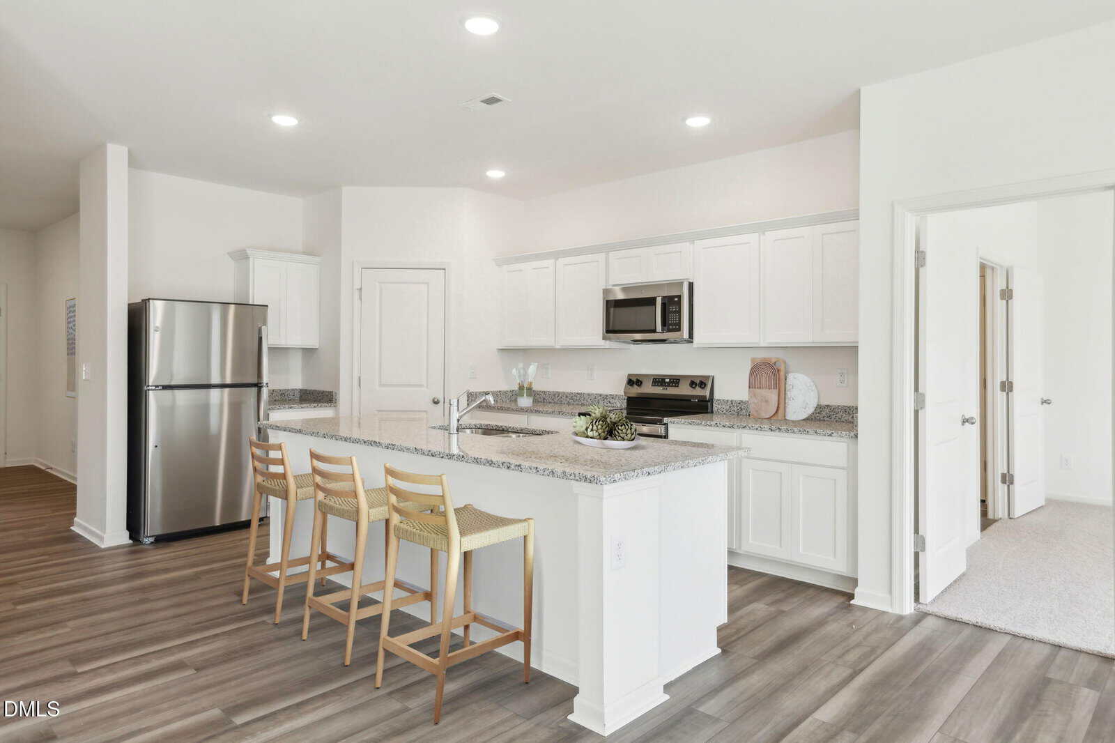 Modern kitchen with white cabinets, granite countertops, stainless steel appliances, and an island with three wooden barstools. Light wood flooring extends throughout, and doors lead to adjacent rooms. Bright and clean design.