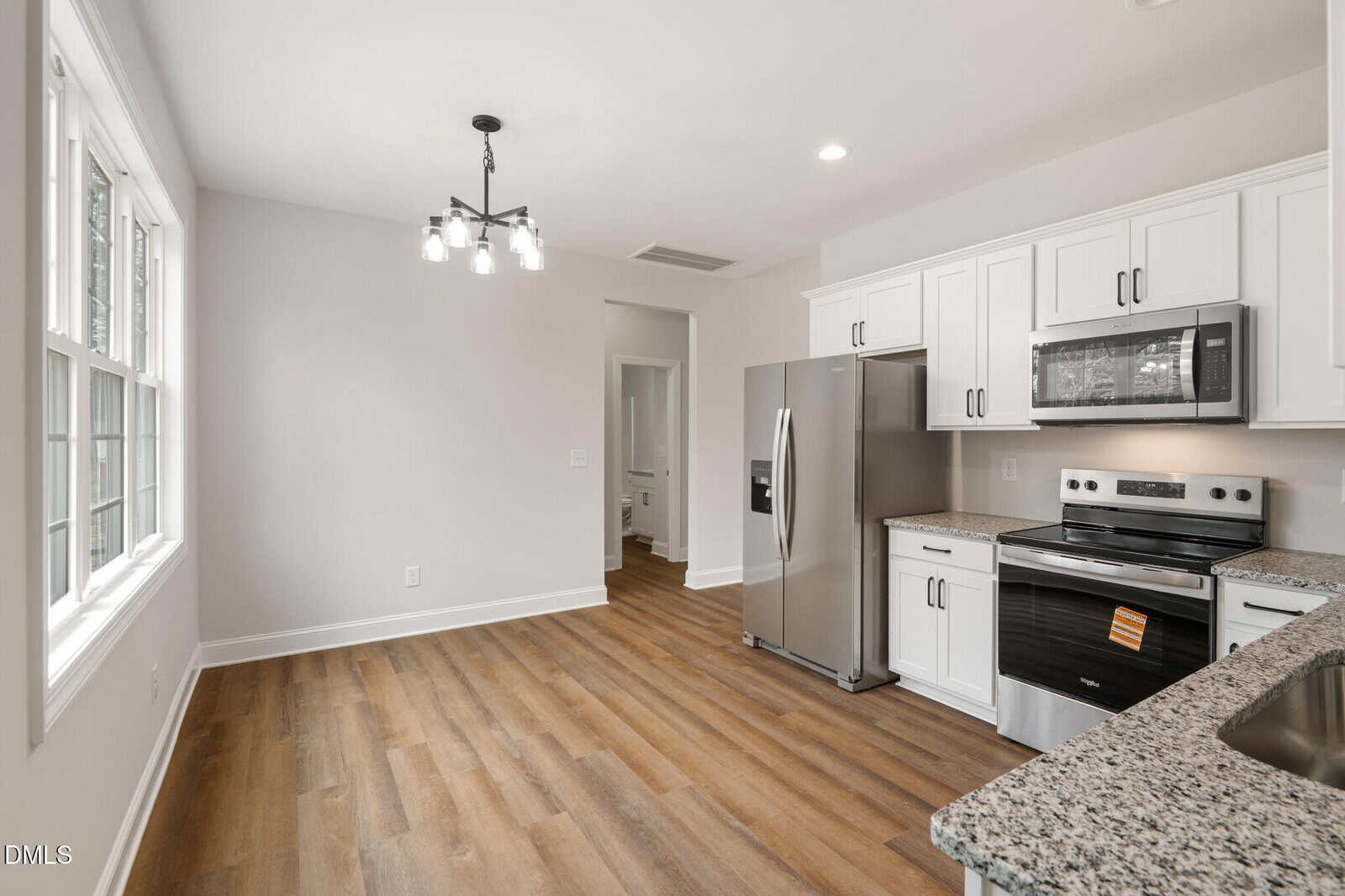 Modern kitchen with white cabinets, stainless steel appliances, granite countertops, and wood-style flooring. Large window lets in natural light. The space includes a dining area and an overhead light fixture.