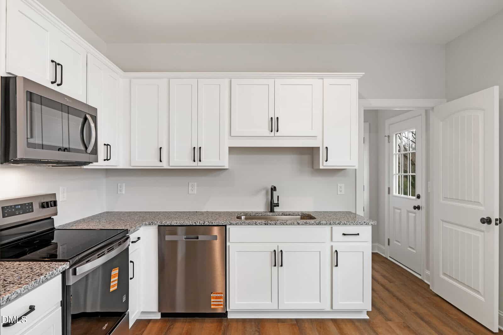Modern kitchen with white cabinets, stainless steel appliances, granite countertops, a built-in microwave, and wood flooring. There is a sink under cabinetry and a white door with glass panes leading outside.
