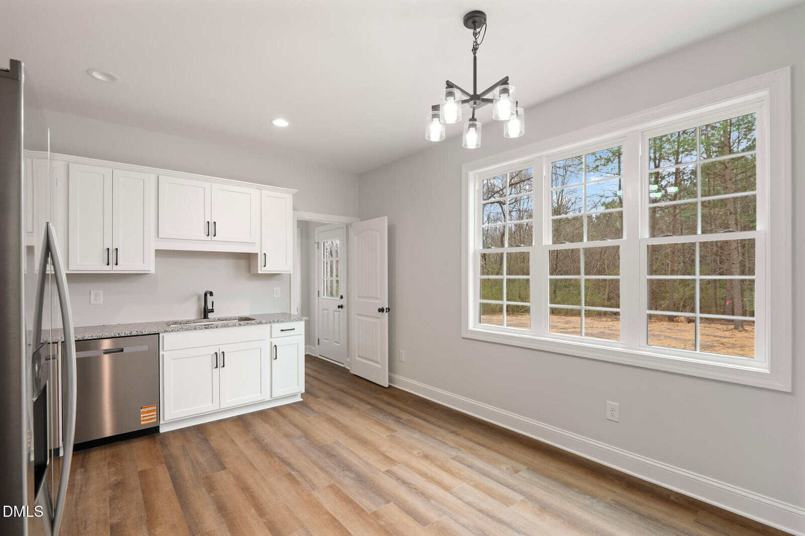 A bright kitchen with white cabinets, stainless steel appliances, wood flooring, a modern light fixture, and a large window showing a view of trees and grass outside.