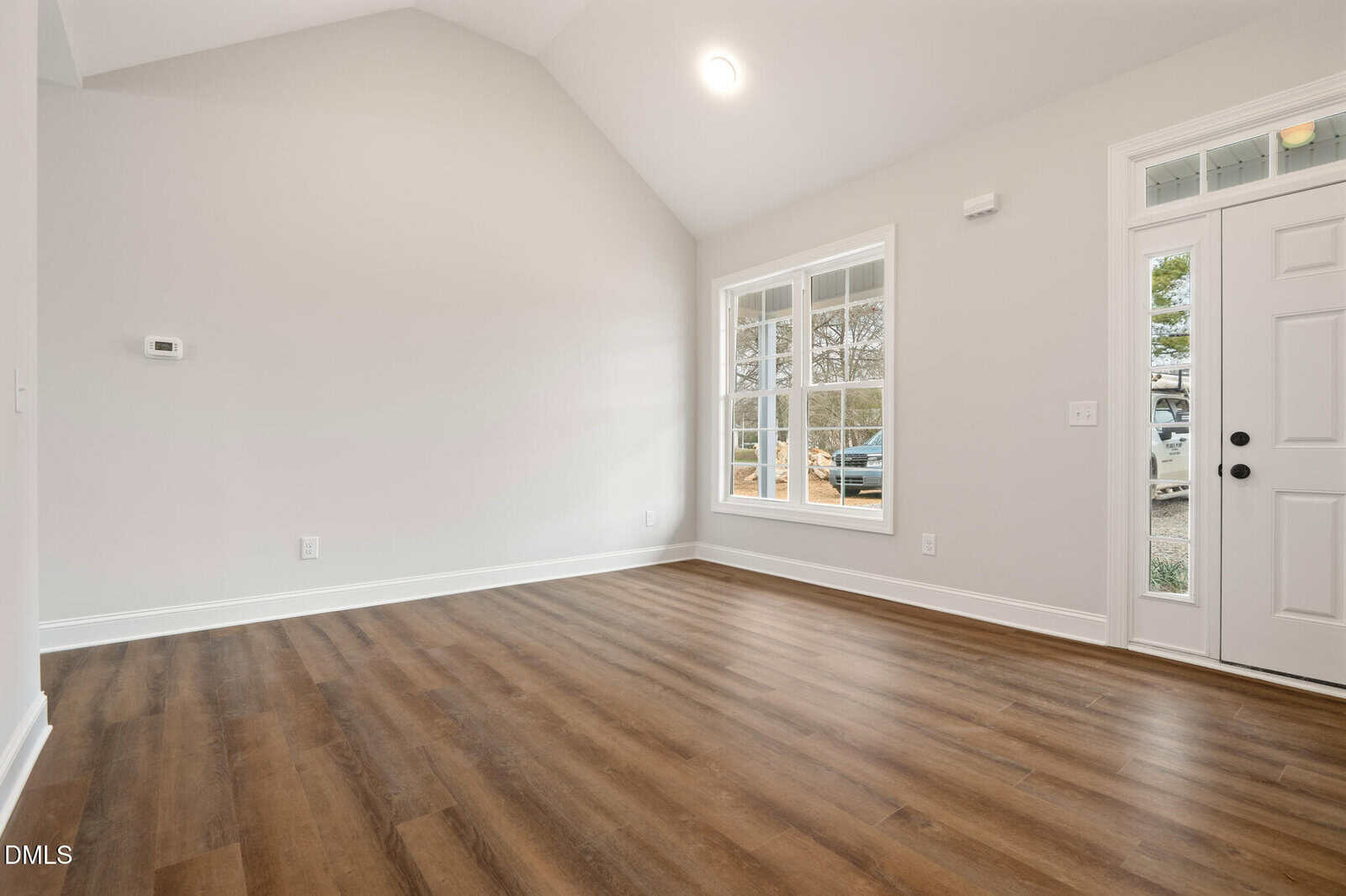 A bright, empty room with vaulted ceilings, light-colored walls, large front window, and wood-look flooring. A white front door with glass panels is to the right, letting in natural light.