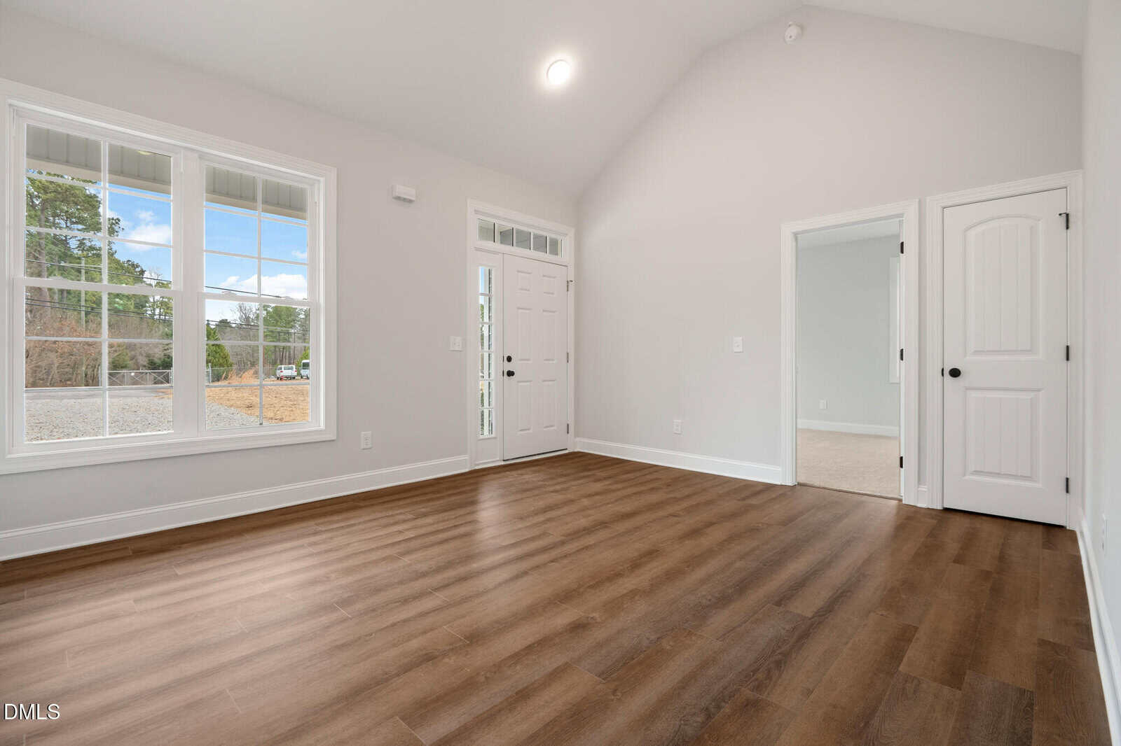 A bright, empty room with light gray walls, large front windows, wood-style flooring, a white front door with glass panels, and two interior doors leading to other rooms.