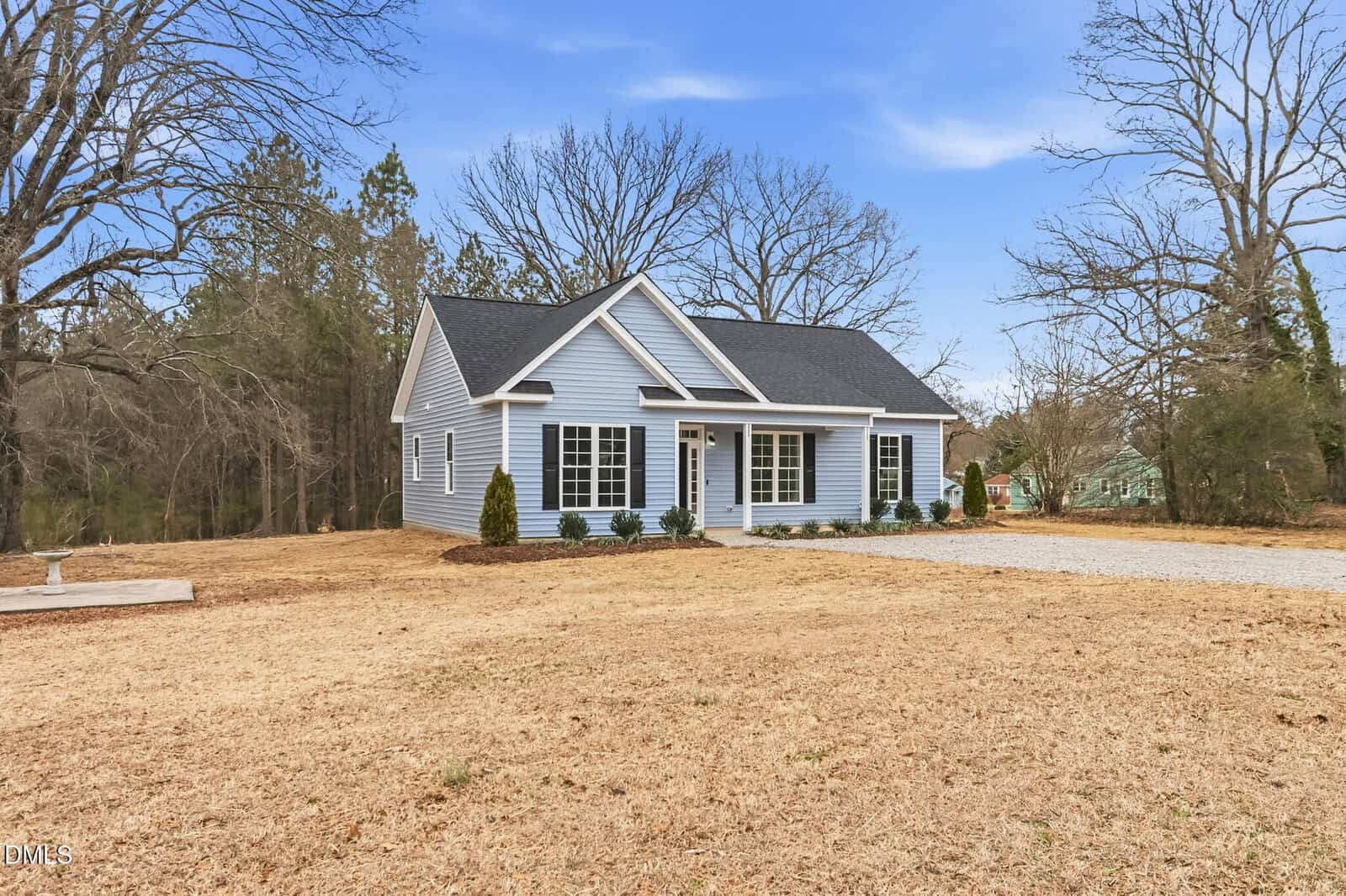 A small, single-story light blue house with white trim and black shutters sits on a large lawn with bare trees and a gravel driveway under a clear sky.