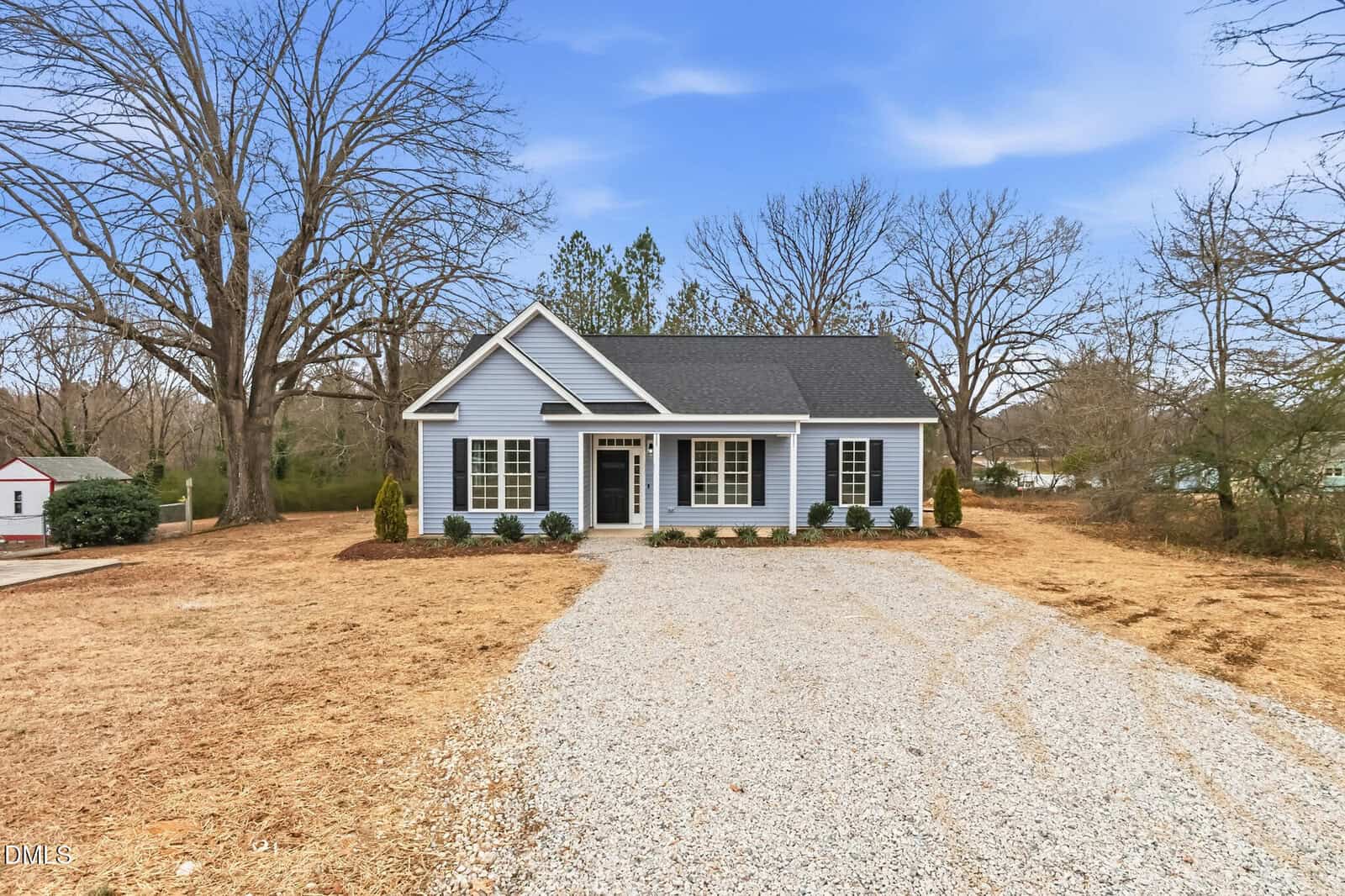 A small, single-story blue house with white trim sits at the end of a gravel driveway, surrounded by dry grass and leafless trees under a clear blue sky.