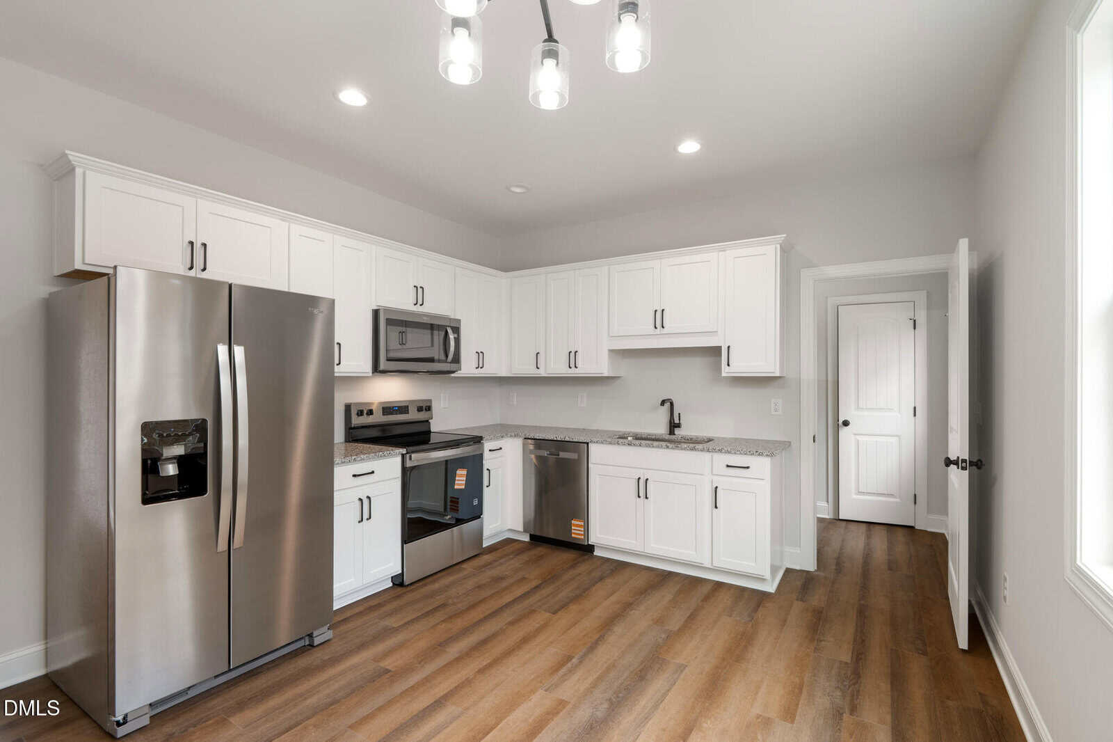 Modern kitchen with white cabinets, stainless steel appliances, granite countertops, wood flooring, and a ceiling light fixture. There is a window on the right and a door at the back of the room.