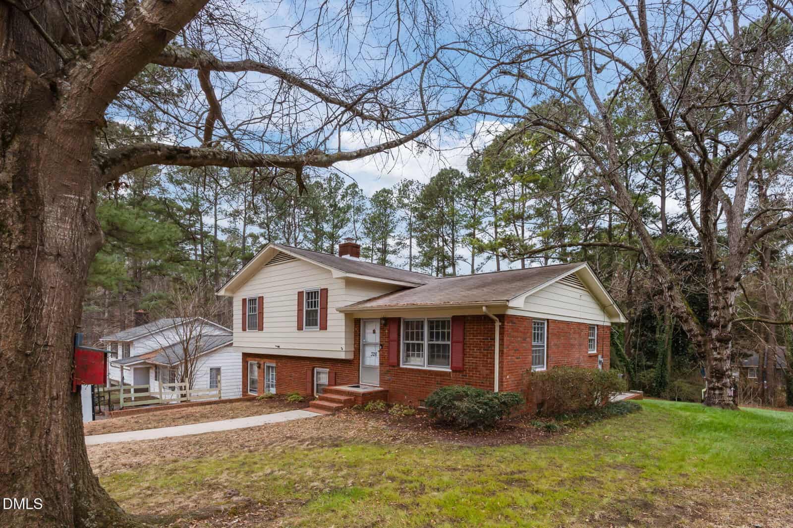 A split-level brick and siding house with a red door sits on a grassy lot at 328 White Oak Drive, Henderson, bordered by trees and featuring a driveway, front steps, and shrubbery near the entrance under a partly cloudy sky.