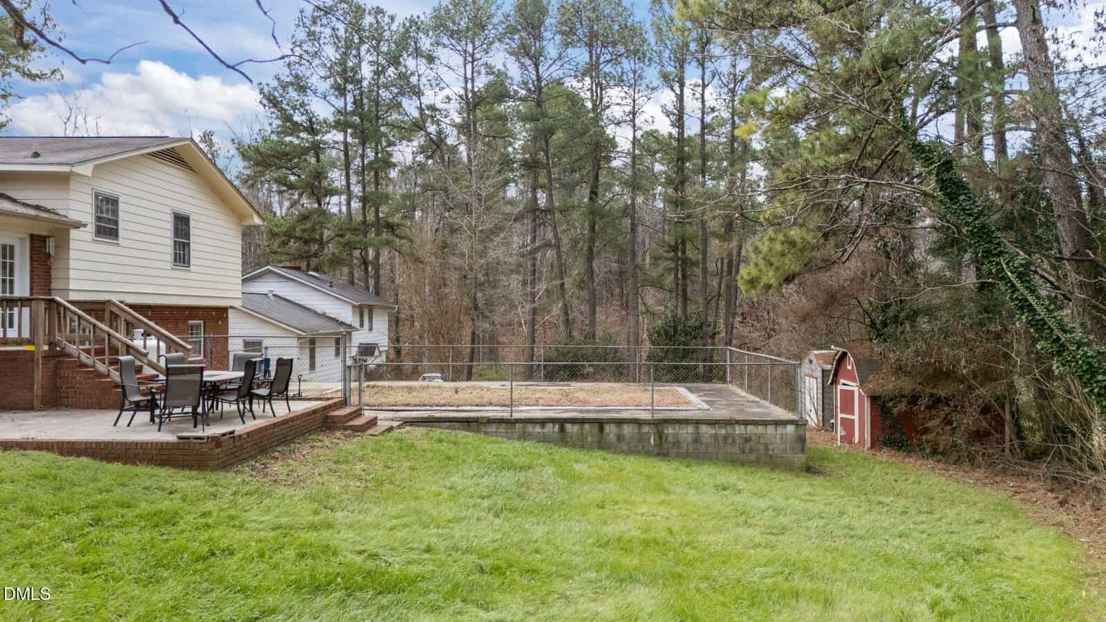 A backyard with a grassy lawn, a raised patio with outdoor dining furniture, a fenced area with bare ground, a small red shed, and tall trees in the background.