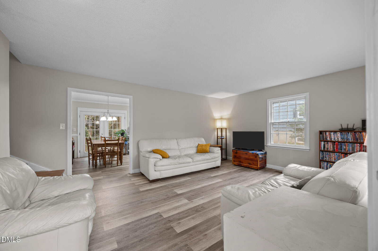 A spacious living room with light wood flooring, white leather sofas, a wooden TV stand with a flat-screen TV, a bookshelf, and large windows letting in natural light. Dining room visible through a doorway.