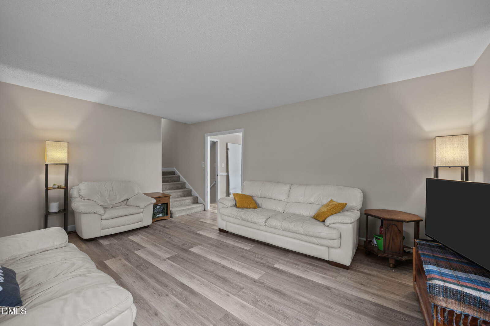 A living room with light wood flooring, beige walls, two white sofas with yellow cushions, two tall lamps, a TV on a wooden stand, and a staircase leading upstairs.