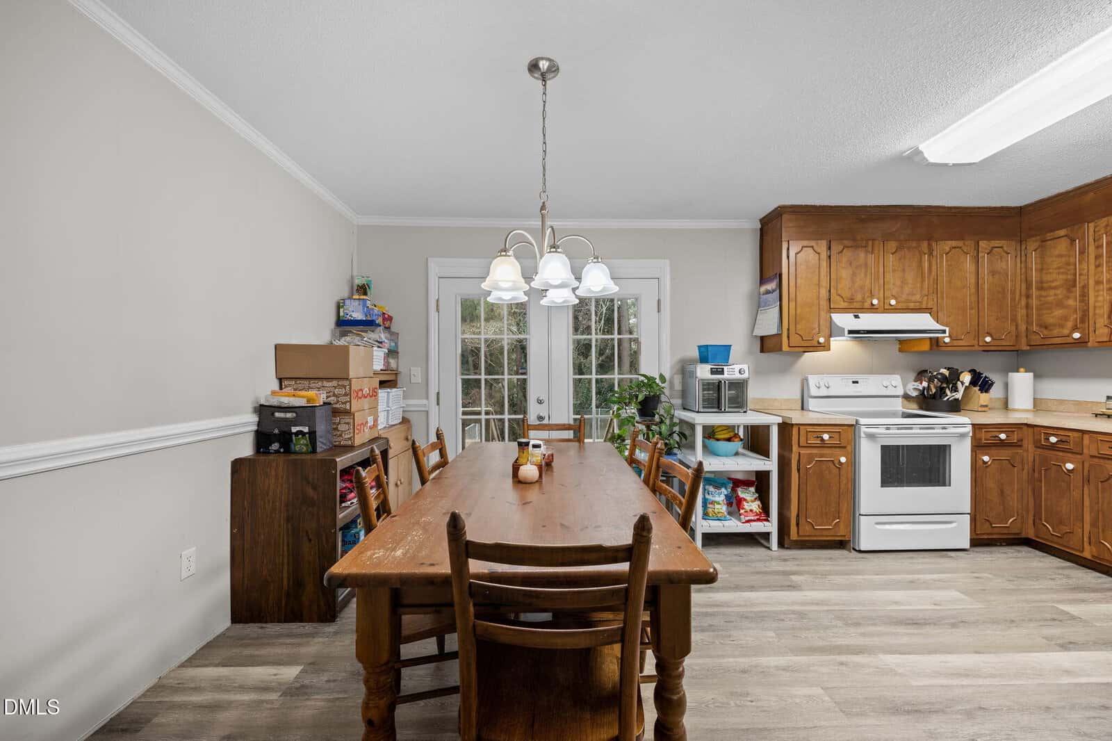 A kitchen and dining area with a wooden table and six chairs, a hanging light fixture, and wooden cabinets. The space includes a white stove, microwave, and glass doors leading outside. Board games are stacked on a side cabinet.