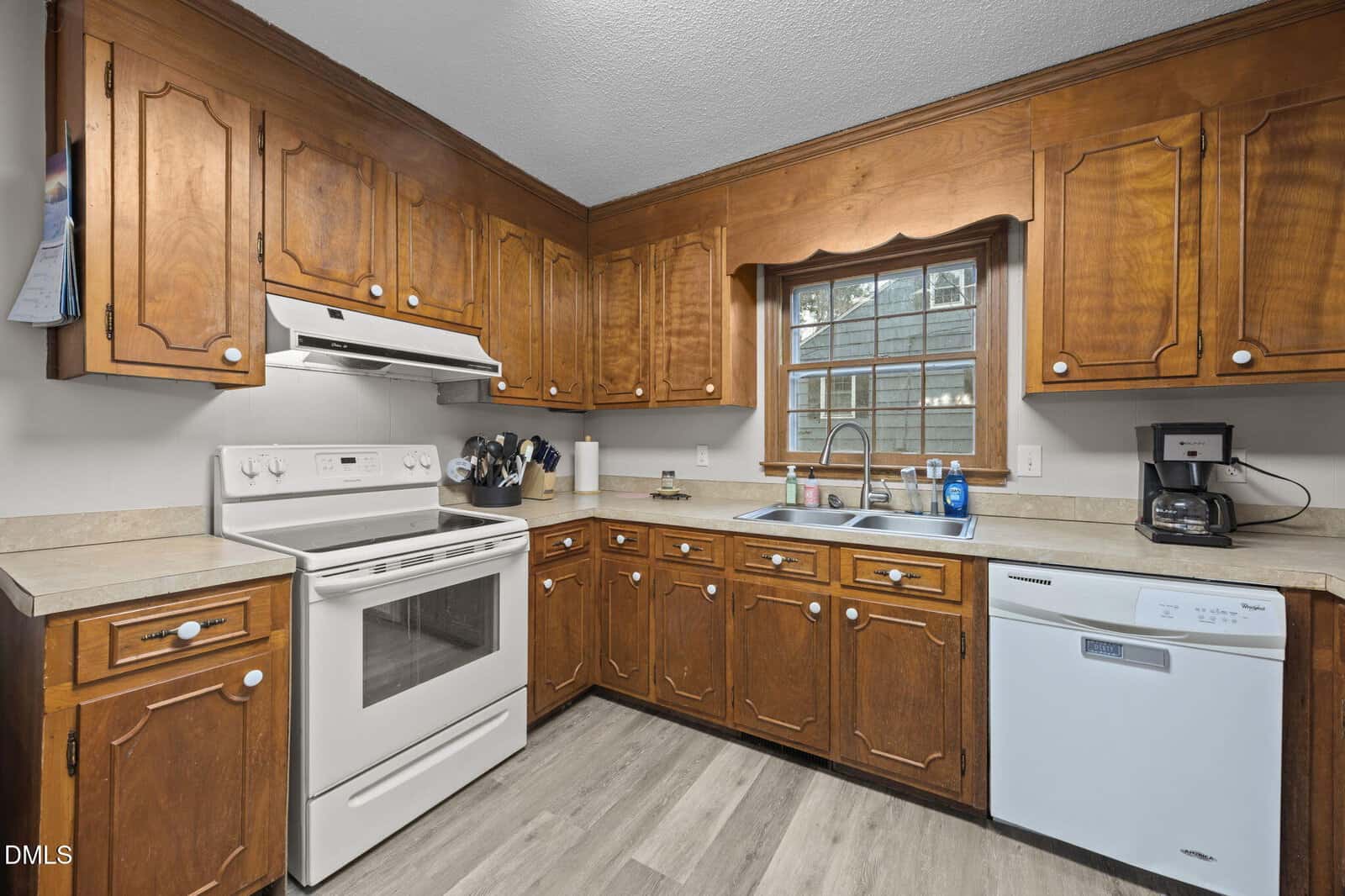 A kitchen with wood cabinets, white appliances including a stove and dishwasher, a double sink under a window, a coffee maker, and utensils on the counter. The floor is light-colored wood.