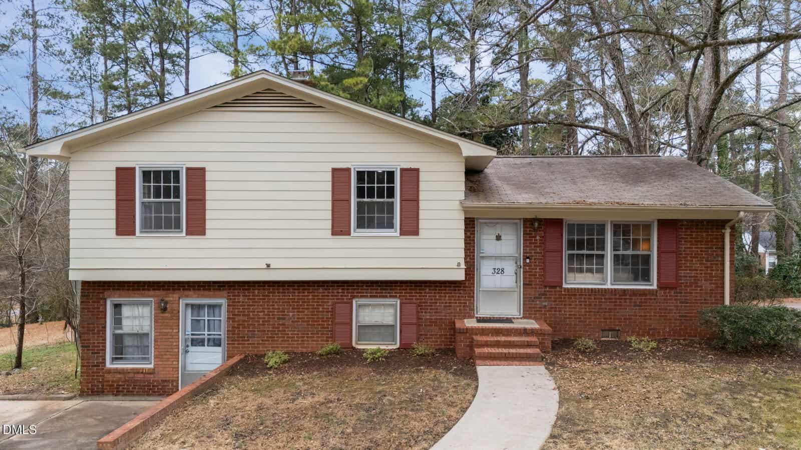 A split-level house with red brick on the lower half and beige siding on the upper half, red shutters, a white front door, and a small front staircase. Tall trees stand in the background.