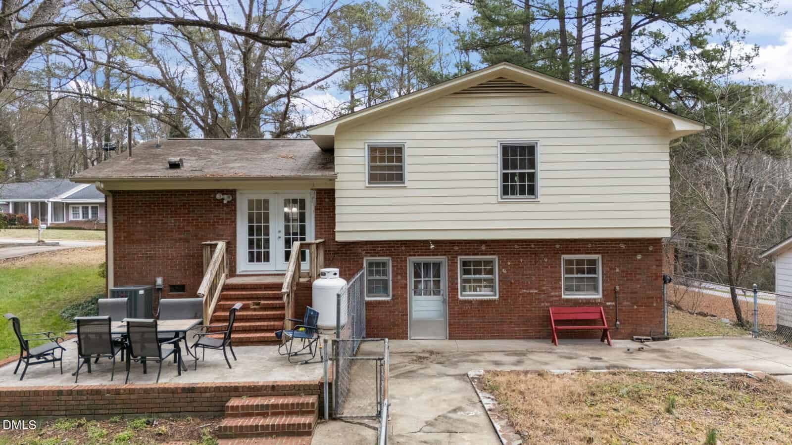 A two-story brick and siding house with a raised wooden deck, outdoor dining set, red bench, and fenced patio area, surrounded by trees and neighboring homes.