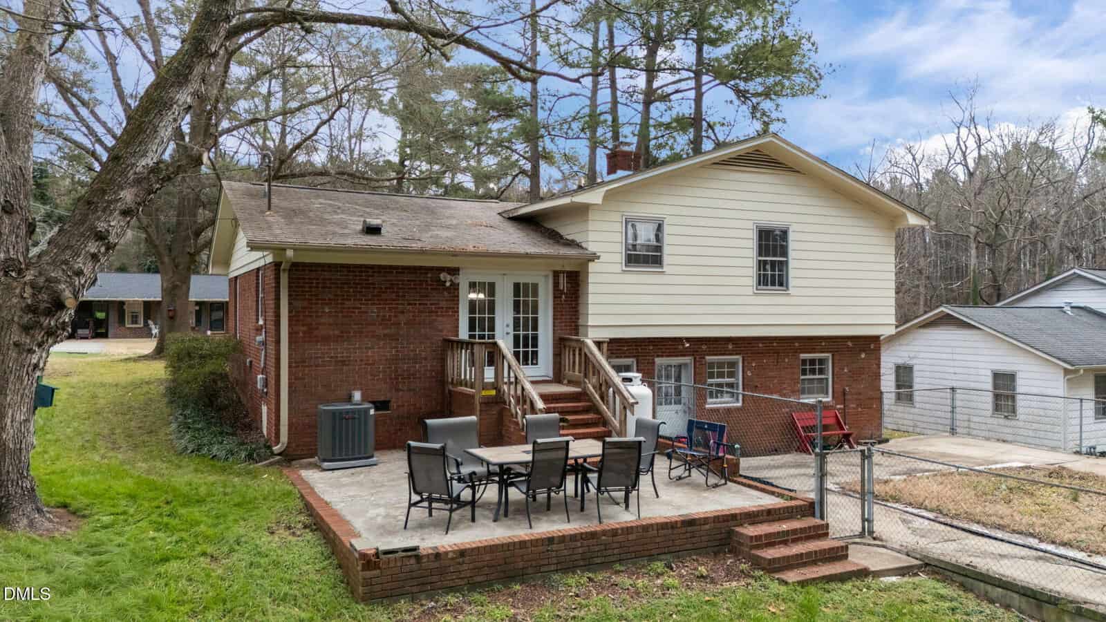A two-story brick house with a patio featuring outdoor dining furniture, surrounded by grass, trees, and neighboring homes under a partly cloudy sky.