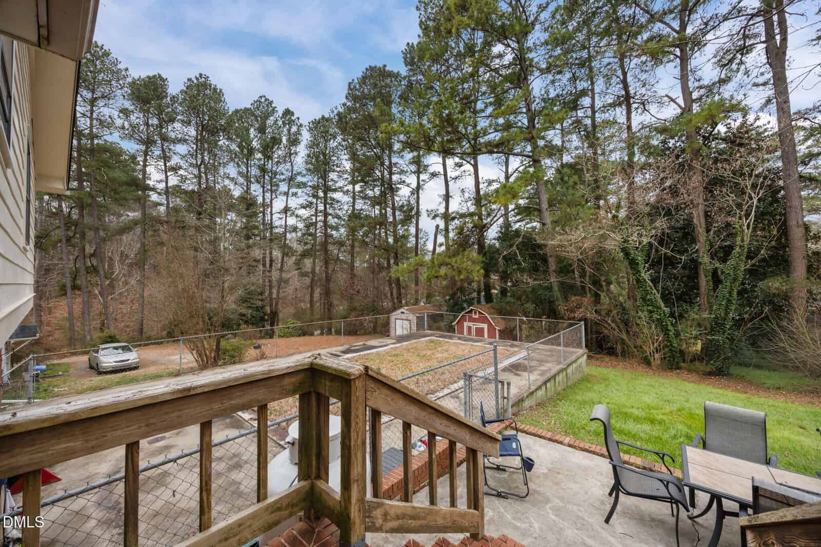 View from a wooden deck overlooking a fenced-in, empty rectangular pool, patio furniture, grassy yard, two red sheds, a parked car, and tall pine trees in the background.