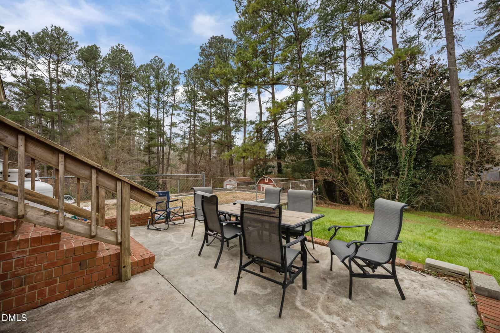 Outdoor patio with a table and six chairs on concrete, next to brick steps. Surrounded by green grass, trees, a shed, and partially cloudy blue sky in the background.
