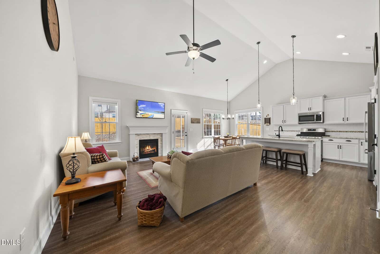 Open-concept living room and kitchen with vaulted ceiling, wood floors, beige sofas, fireplace, wall-mounted TV, dining table by windows, and white cabinets with stainless steel appliances and a large island.
