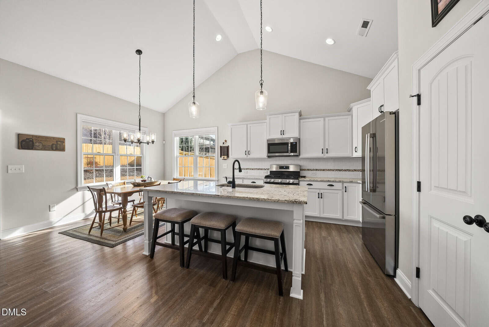 Modern kitchen with white cabinets, granite countertops, stainless steel appliances, and an island with three stools. Dining area with table and chairs by large windows. Wood flooring and pendant lighting overhead.