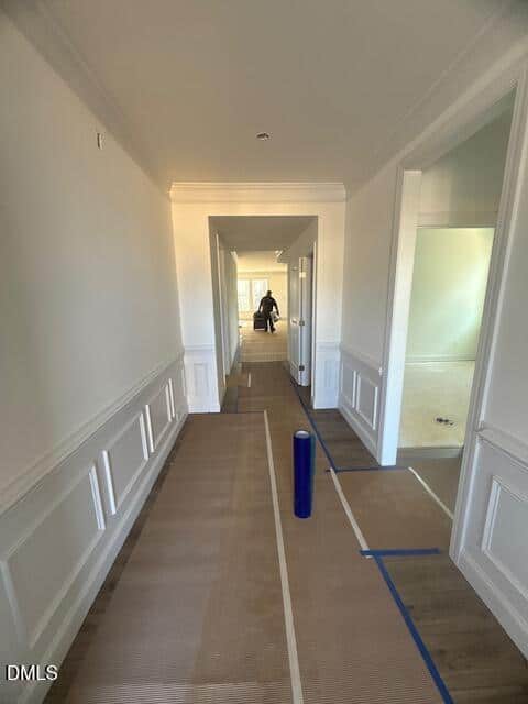 A hallway in a house with white walls and wainscoting, protective paper covering the floor, blue tape along the edges, and a person working in the distance near a doorway.
