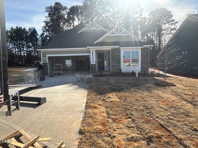 A single-story house under construction with a “SOLD” sign in the window. Construction materials and equipment are on the driveway, and a worker is present. The yard is patchy with brown grass. Trees are in the background.