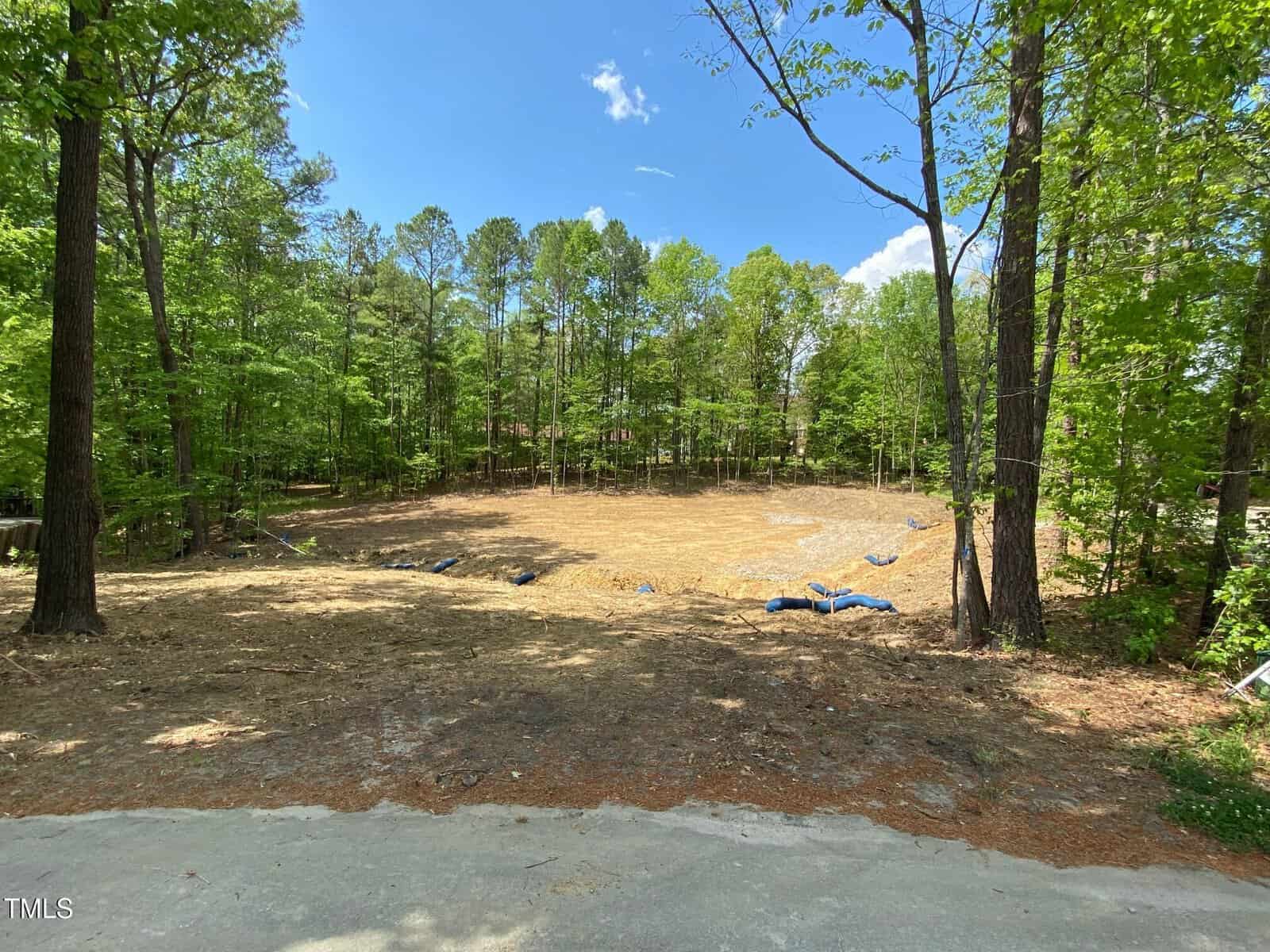 A cleared, dirt-covered area in a forested landscape, bordered by tall trees under a blue sky with scattered clouds. Some rolled-up tarps and straw cover parts of the ground.