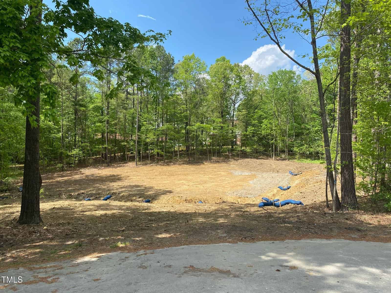 A cleared, dirt lot surrounded by tall green trees on a sunny day, with blue sandbags scattered along the ground and a patch of pavement visible in the foreground.