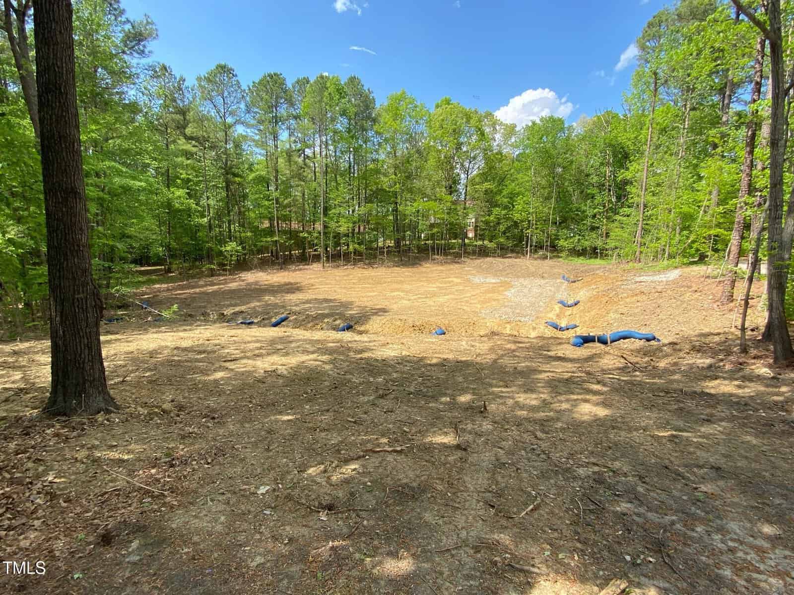 A cleared, dirt lot surrounded by tall, green trees on a sunny day, with erosion control barriers and straw covering parts of the ground.