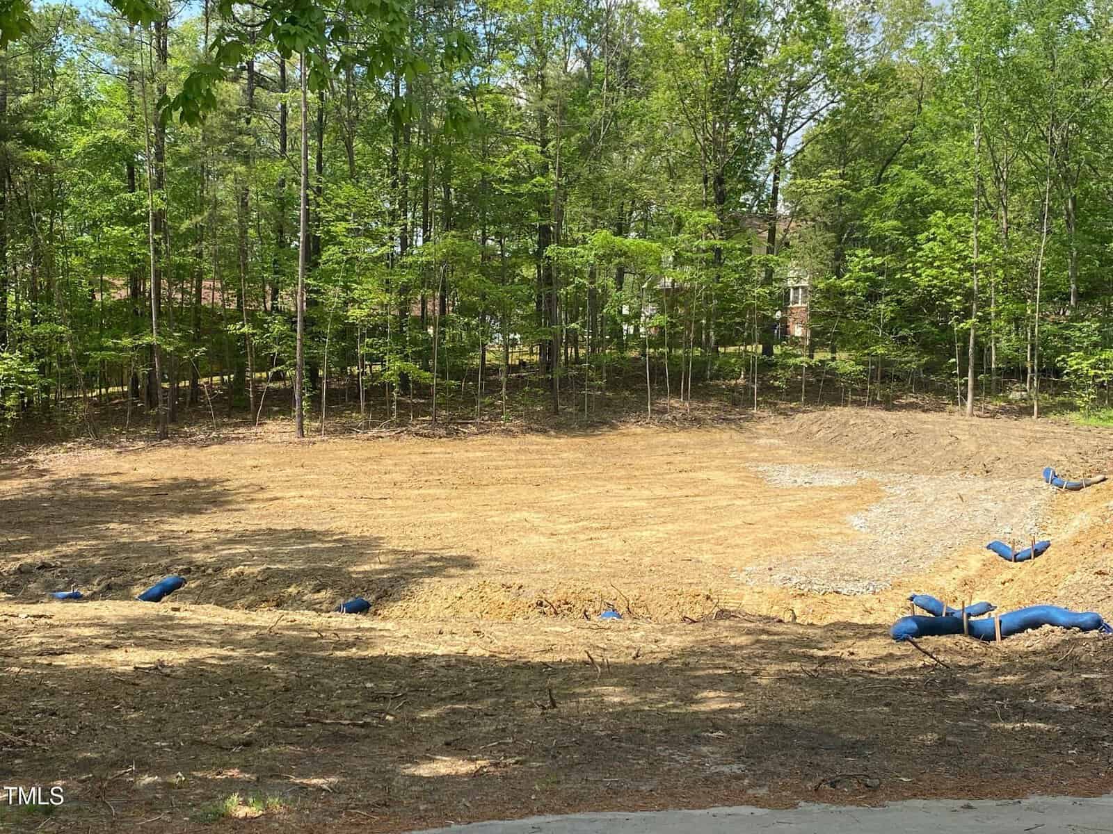 A cleared and leveled patch of land with some construction materials and small barriers, surrounded by a dense forest of green trees on a sunny day.