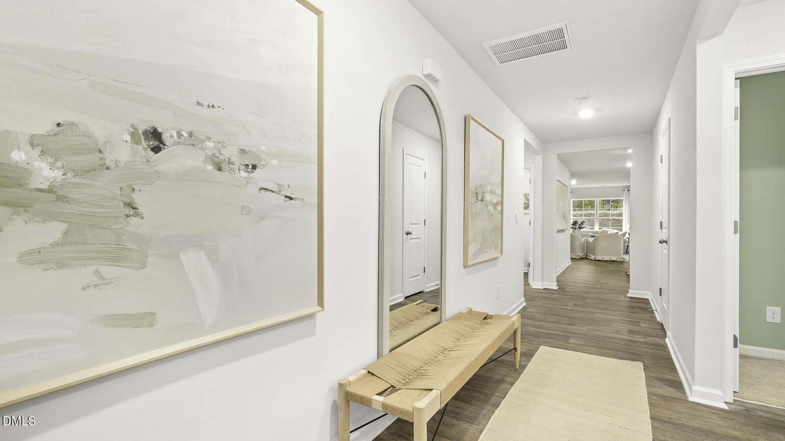 A bright, modern hallway with light walls, wood flooring, a large abstract painting, a woven bench, a round mirror, and a beige rug. The hallway leads to a sunlit living space with large windows.