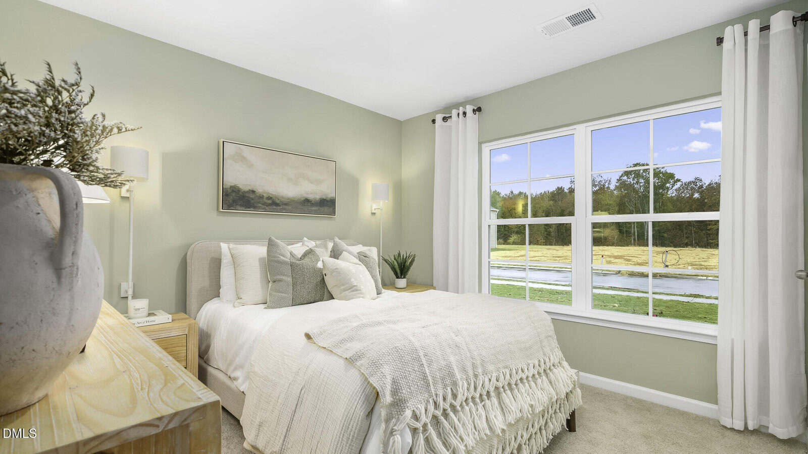 Bright bedroom with a beige bed, textured bedding, and decorative pillows. A large window lets in natural light, showing an outdoor view. Artwork and a plant decorate the soft green walls. A wooden nightstand sits nearby.